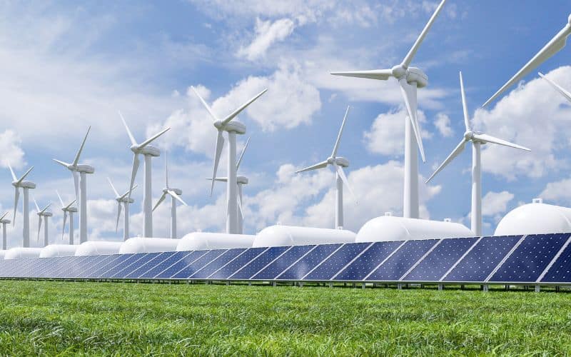 A renewable energy farm with rows of solar panels in the foreground and multiple wind turbines under a cloudy sky.