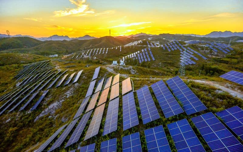 Aerial view of a vast solar farm with rows of solar panels set in a hilly landscape at sunset, emphasizing renewable energy.